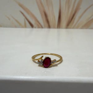Peut inclure: Une bague en or ornée d'une pierre précieuse rouge foncé de taille ovale au centre, entourée de petites pierres claires de chaque côté. L'anneau est fin et poli, présentant un sertissage de style bypass qui maintient les pierres en place.