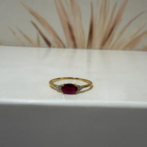 Peut inclure: Une bague en or ornée d'une pierre précieuse rouge foncé de taille ovale au centre. La pierre est flanquée de chaque côté de deux petites pierres d'accent claires et scintillantes serties sur le fin anneau en or poli et brillant.