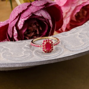 Peut inclure: Une bague en or avec une pierre précieuse rouge rectangulaire centrale entourée d'un halo de petites pierres rouges, avec d'autres pierres rouges serties le long du fin anneau en or. La bague est posée sur une surface grise incurvée.
