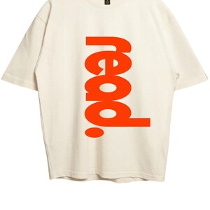 Puede incluir: Una camiseta de manga corta color blanco roto con la palabra read. impresa en letras grandes, negritas y verticales de color naranja en el centro. La prenda tiene cuello redondo y una etiqueta de la marca Gildan en el interior del cuello.