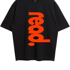 Puede incluir: Una camiseta negra de manga corta con la palabra "read." impresa verticalmente en letras grandes, negritas y de color naranja brillante en el centro. La prenda tiene un cuello redondo y una etiqueta de la marca Gildan visible en el interior.