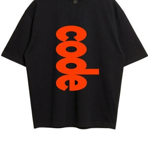 Puede incluir: Una camiseta negra de manga corta con la palabra code impresa verticalmente en letras de color naranja brillante en el centro. La camiseta tiene un cuello redondo y una etiqueta de la marca Gildan es visible en el interior del cuello.