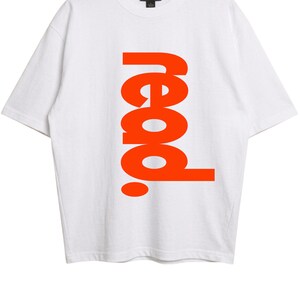 Puede incluir: Una camiseta blanca de manga corta con la palabra "read." impresa verticalmente en letras grandes, negritas y de color naranja brillante en el centro. La prenda tiene un cuello redondo y una etiqueta de la marca Gildan visible en el interior.