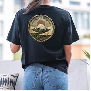 Puede incluir: Una camiseta negra con un gráfico circular en la espalda. El diseño muestra el monte Fuji con un sol rojo, nubes y el texto "Mount Fuji" y "3,776 m" con un acabado metálico desgastado y un borde envejecido.