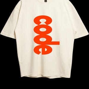 Puede incluir: Una camiseta de manga corta de color crema con la palabra code impresa en una fuente naranja brillante y negrita verticalmente en el centro del pecho. La prenda tiene un cuello redondo y una etiqueta de la marca Gildan visible en el interior.