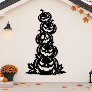May include: A black metal wall art piece featuring a vertical stack of five carved jack-o'-lanterns with jagged, triangular eyes and toothy grins, accented by stylised leaf shapes at the base, mounted on a light-coloured exterior wall.