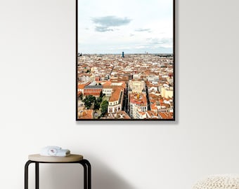 May include: A framed vertical wall art print featuring a high-angle panoramic view of a dense city landscape with numerous buildings topped with terracotta-coloured roofs under a bright, overcast sky with scattered clouds.