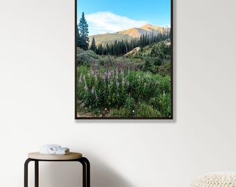 May include: A framed vertical wall art print featuring a landscape photograph of a mountain range under a clear blue sky, with a foreground of green foliage and purple wildflowers, and a dense forest of pine trees covering the mountain slopes.