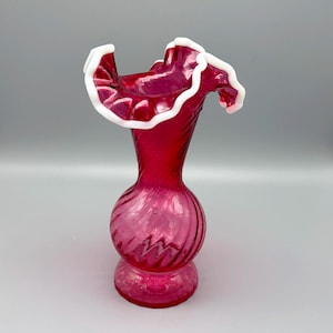 May include: A cranberry red glass vase featuring a bulbous base and a slender neck with a spiraling ribbed texture. The top opening is wide and flared with a distinct, wavy, ruffled edge finished in an opaque white glass trim.