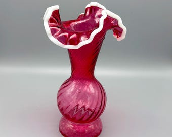 Könnte beinhalten: Eine cranberryrote Glasvase mit bauchigem Fuß und schlankem Hals mit spiralförmiger Rippenstruktur. Die obere Öffnung ist breit und ausgestellt mit einem markanten, welligen, gerüschten Rand, der mit einem opaken weißen Glasbesatz versehen ist.