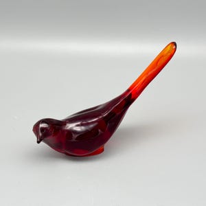 May include: A stylized bird figurine crafted from smooth, translucent glass. The body features a deep, saturated ruby red hue that transitions into a vibrant, bright orange color along the elongated, pointed tail section, creating a gradient effect.