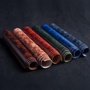 May include: Six rolls of leather arranged in a row, featuring a mottled, distressed texture. The colors include dark brown, light brown, reddish-brown, forest green, royal blue, and navy blue, each showing a smooth interior surface.