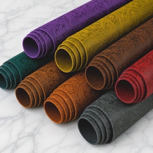 May include: A collection of seven rolled sheets of material in various colors including purple, mustard yellow, dark brown, teal, light brown, red, and charcoal gray. Each sheet features a consistent embossed paisley pattern across the surface.