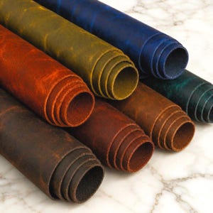 Op de afbeelding: Een collectie opgerolde leren vellen in diverse kleuren, waaronder diepblauw, olijfgroen, feloranje, donkerbruin en groenblauw. Elke rol heeft een mat, getextureerd oppervlak met zichtbare nerfpatronen en natuurlijke variaties in de kleur.