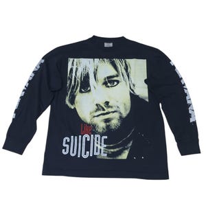 Puede incluir: Una camiseta negra de manga larga con una gran impresión fotográfica centrada de Kurt Cobain. La palabra "Like" está impresa en rojo sobre la palabra "SUICIDE" en letras blancas. Las mangas presentan texto blanco a lo largo.