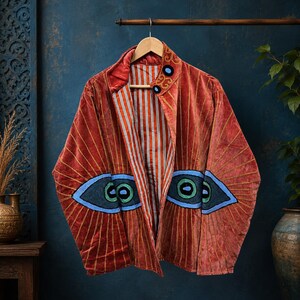 Könnte beinhalten: Eine rostfarbene Samtjacke mit gestickten Augenmotiven auf den Vorderteilen. Die Jacke hat einen Stehkragen, lange Ärmel und ein vertikal gestreiftes Innenfutter in Orange, Weiß und Schwarz. Goldfarbene Nähte strahlen von den Augen aus.
