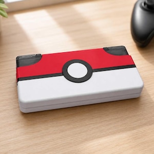 May include: A rectangular handheld gaming console case featuring a red top half and a white bottom half, separated by a horizontal black stripe. A central circular button design with a white center and black border is positioned on the front surface.