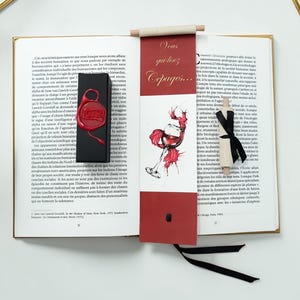 Op de afbeelding: Een open boek met een zwarte rechthoekige doos met een rood waszegel, een verticale bladwijzer met een rood bovengedeelte met de tekst "Vous qui lisez Cepages..." en een wijnglasillustratie, en een kleine rol vastgebonden met een zwart lint.