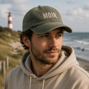 Könnte beinhalten: Eine olivgrüne Baseballkappe mit dem Wort MOIN. in weißer Stickerei in Großbuchstaben auf der Vorderseite. Die Kappe verfügt über eine strukturierte Krone, einen gebogenen Schirm und einen Stoffriemen mit Metallschnalle zur Größenanpassung.
