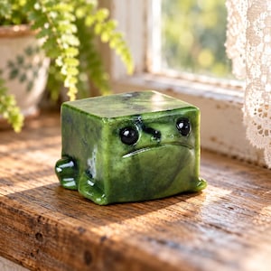 May include: A cube-shaped green stone figurine resembling a frog with a smooth, polished surface, dark marbled veining, and two small, round, black inset eyes. The object features a flat top and small, rounded protrusions for feet.
