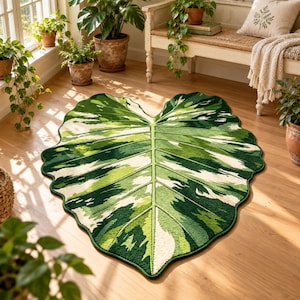 May include: A large, leaf-shaped area rug featuring a variegated pattern of dark green, light green, and cream colors. The rug mimics the appearance of a tropical plant leaf with a prominent central vein and smaller lateral veins, with a scalloped edge.