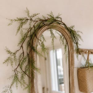 Può includere: Uno specchio ad arco con una cornice in legno marrone chiaro dall'aspetto vissuto, drappeggiato con una lunga ghirlanda artificiale di cedro con sottili rami verdi testurizzati che si estendono sopra la parte superiore e lungo il lato sinistro.
