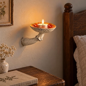 Peut inclure: Un bougeoir mural en forme de champignon avec un pied texturé blanc cassé et un chapeau rouge à pois blancs. Le dessus du chapeau est concave et accueille une petite bougie chauffe-plat allumée avec une flamme chaude et lumineuse.