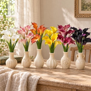 Puede incluir: Seis pequeños jarrones de cerámica, cada uno con un ramo de calas artificiales en colores distintos: blanco, rosa claro, naranja, amarillo, magenta y morado oscuro. Los jarrones presentan texturas variadas, incluyendo diseños acanalados y en relieve.