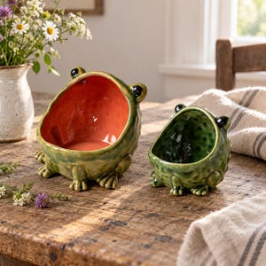 May include: Two ceramic frog-shaped bowls with textured green exteriors and black eyes. The larger bowl features a smooth, bright orange interior, while the smaller bowl has a dark green interior. Both bowls have four small feet at the base.