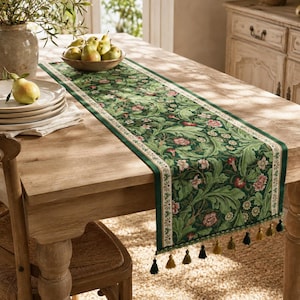 Puede incluir: Un camino de mesa rectangular con fondo verde oscuro y un denso patrón botánico de hojas de acanto, enredaderas y pequeñas flores rosas y blancas. El camino tiene un borde de color crema y borlas decorativas en los extremos.