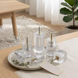 May include: A collection of clear acrylic furniture legs in various shapes, including cylindrical and square designs. Each piece features a metal screw protruding from the top for attachment, displayed on a round ceramic tray.