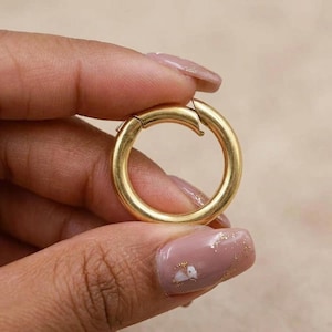 May include: A circular gold-toned metal spring gate ring featuring a smooth, polished finish. The ring has a hinged mechanism that allows the segment to open and close, creating a secure loop for connecting various items together.
