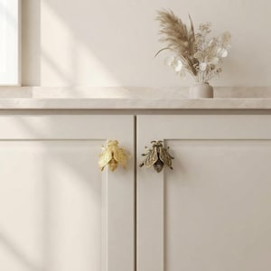 May include: Two bee-shaped cabinet knobs attached to light-colored cabinet doors. One knob is a bright, polished gold finish, while the other features a darker, antique brass finish with detailed wings and body segments.