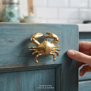 Puede incluir: Un pomo de armario de metal dorado con forma de cangrejo, montado en una puerta de armario de madera de color azul verdoso desgastado. El texto "HANDCRAFTED COASTAL ELEGANCE" está impreso en letras blancas pequeñas en la parte inferior.