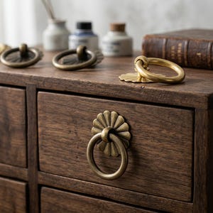May include: A dark brown wooden cabinet featuring multiple drawers, each equipped with a circular brass ring pull attached to a decorative flower-shaped backplate. Additional matching brass ring pulls are placed on the top surface.