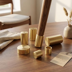 Pode incluir: Uma coleção de capas cilíndricas para pés de móveis em latão maciço de vários tamanhos, com uma capa encaixada num pé de madeira. Uma etiqueta de papel com o texto SOLID BRASS LEGS CAPS está colocada por perto. Os componentes de latão têm um acabamento metálico liso.