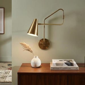 Peut inclure: Une applique murale en laiton dotée d'un abat-jour conique et d'un bras articulé réglable. La lampe présente une plaque murale circulaire et une finition métallique. Elle est placée au-dessus d'une surface en bois avec un vase blanc et des livres.