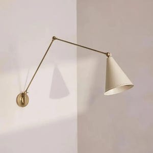 May include: A wall-mounted swing arm lamp featuring a circular brass base, a long adjustable metal arm, and a conical off-white shade. The shade has a perforated edge detail, and the fixture is connected by articulated joints for positioning.