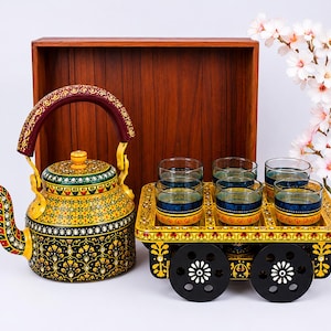 May include: A hand-painted metal tea kettle with a yellow and black floral pattern and a matching cart-shaped serving tray holding six glass tumblers. The tray features black wheels with white floral accents and a wooden back panel.