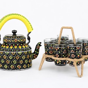 May include: A black metal tea kettle with a bright yellow handle and a hand-painted floral pattern in yellow, red, and green. Beside it sits a matching set of six glass tumblers held in a tan rope-wrapped metal carrier with a central handle.