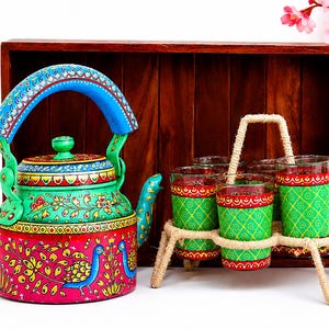 Puede incluir: Una tetera de metal pintada a mano con colores vibrantes rosa, verde y azul con motivos de pavo real, acompañada de un juego de seis vasos a juego en un soporte de yute trenzado, frente a una bandeja rectangular de madera oscura.