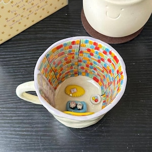 Puede incluir: Una taza de cer&aacute;mica con una escena interior pintada a mano de una biblioteca con estanter&iacute;as coloridas, una alfombra amarilla con un libro, una cama azul para mascotas con dos figuras peque&ntilde;as y un cuenco con bolas de colores.