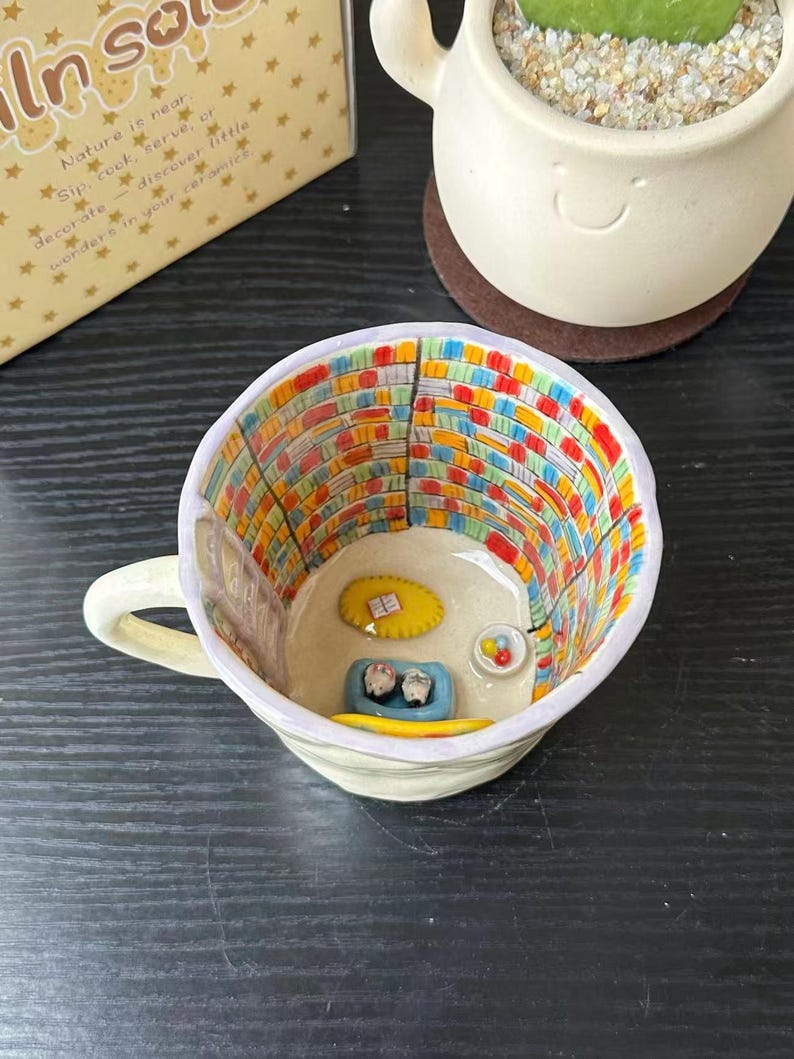 Puede incluir: Una taza de cer&aacute;mica con una escena interior pintada a mano de una biblioteca con estanter&iacute;as coloridas, una alfombra amarilla con un libro, una cama azul para mascotas con dos figuras peque&ntilde;as y un cuenco con bolas de colores.