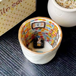 May include: A white ceramic cup featuring a hand-painted interior scene of a library with colourful bookshelves, a ladder, and a small black armchair. The exterior is plain white with a brown rim. The packaging box displays the text "Sip in Solo".
