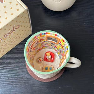 Może przedstawiać: Ceramiczny kubek z ręcznie malowaną sceną wnętrza biblioteki z kolorowymi regałami, małym czerwonym fotelem z figurką misia, dywanikiem i stolikiem. Zewnętrzna strona jest kremowa z jasnozielonym brzegiem i uchwytem.