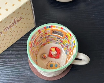 Puede incluir: Una taza de cerámica con una escena interior pintada a mano de una biblioteca con estanterías coloridas, un pequeño sillón rojo con una figura de oso, una alfombra y una mesa auxiliar. El exterior es de color crema con un borde verde claro.