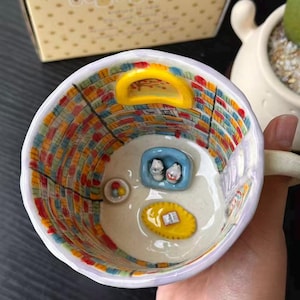Puede incluir: Una taza de cer&aacute;mica con un interior pintado a mano que muestra una escena de biblioteca, incluyendo estanter&iacute;as coloridas, una puerta arqueada amarilla, un sof&aacute; azul con dos figuras, una alfombra amarilla con un libro y un cuenco con esferas.