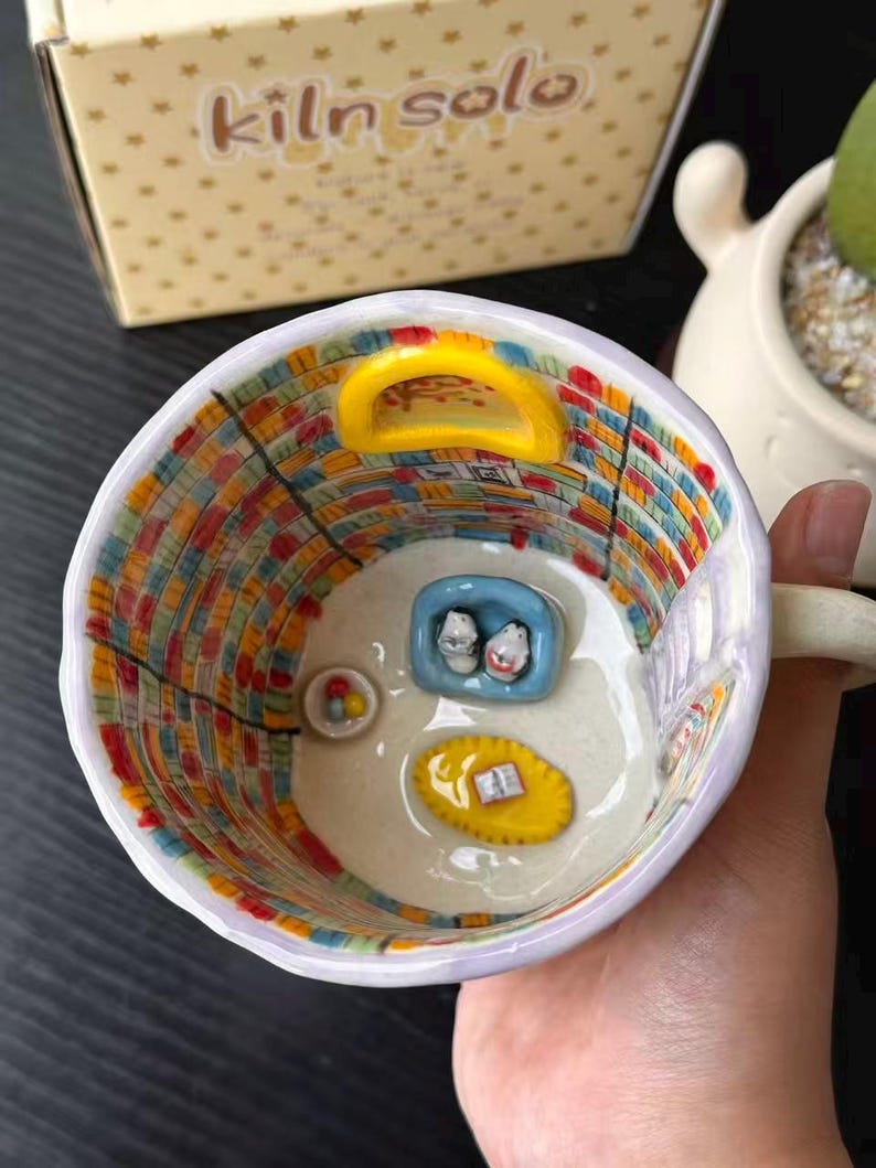 Puede incluir: Una taza de cer&aacute;mica con un interior pintado a mano que muestra una escena de biblioteca, incluyendo estanter&iacute;as coloridas, una puerta arqueada amarilla, un sof&aacute; azul con dos figuras, una alfombra amarilla con un libro y un cuenco con esferas.
