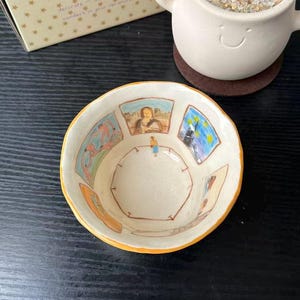 Puede incluir: Un cuenco de cerámica color crema con borde amarillo, que presenta ilustraciones pintadas a mano de famosas pinturas de galerías de arte en las paredes interiores y una pequeña figura en el centro, simulando el interior de un museo.