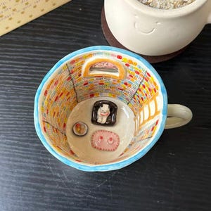 Könnte beinhalten: Ein Keramikbecher mit hellblauem Rand und einem Innenraum, der wie ein Zimmer mit Bücherregalen, einer Leiter und einem Fenster bemalt ist. Im Inneren sitzt ein kleiner weißer Bären-Anhänger in einem dunklen Sessel neben einem rosa Teppich.
